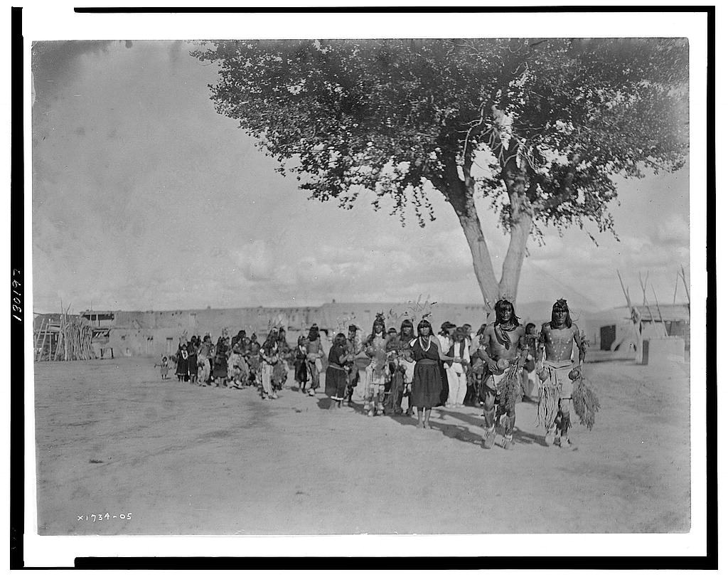 A large group of Pueblo people in traditional ceremonial regalia gather beneath a spreading tree in an arid landscape, with two prominent figures in elaborate