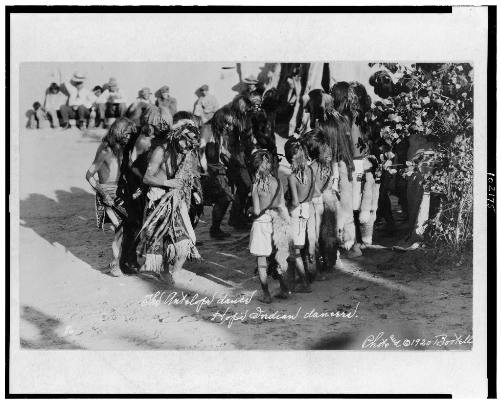 Four shirtless Hopi dancers in geometric-patterned kilts perform energetically while a seated audience watches in bright sunlight.