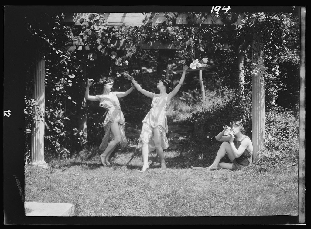 Three women in white tunics leap and reach upward on a grass lawn framed by classical columns, 1911.