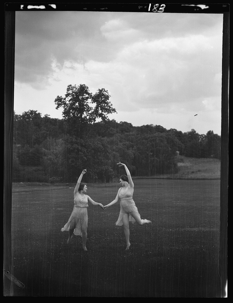 Two barefoot dancers in draped tunics hold hands while turning in an open field, with woodland and a large tree behind them under a cloudy sky.