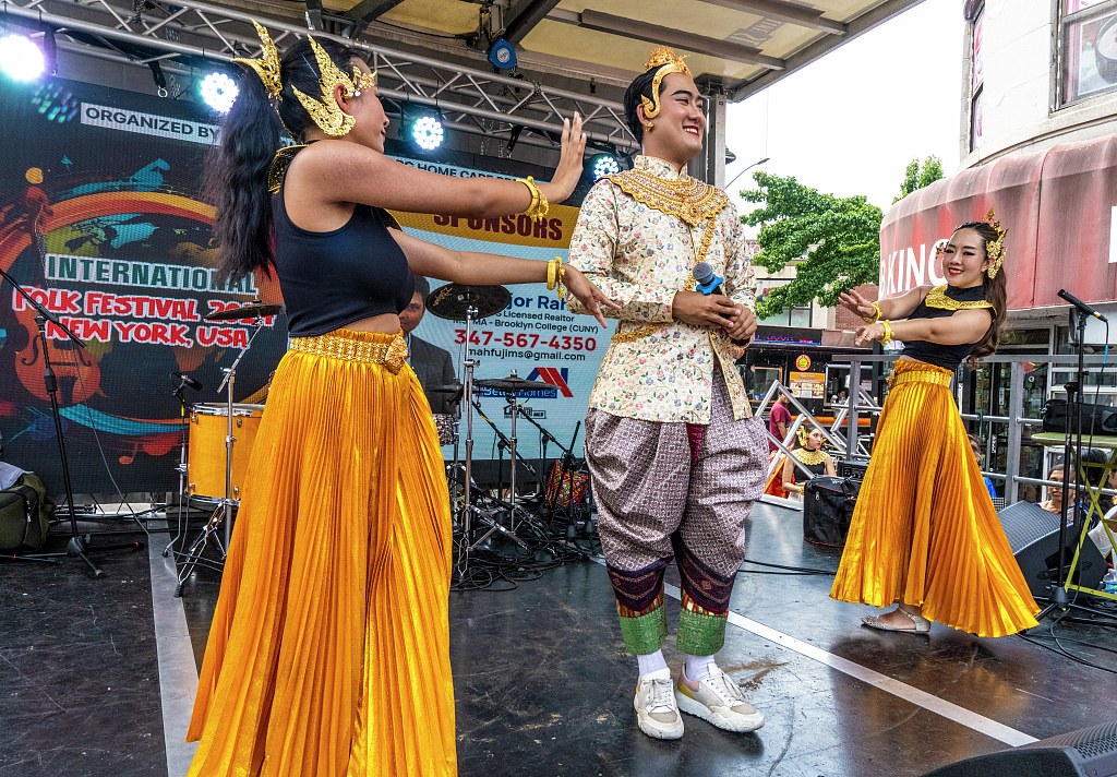 Three performers in Thai silk costumes at outdoor stage: male singer in center with arms raised, two female dancers symmetrically positioned on either side.