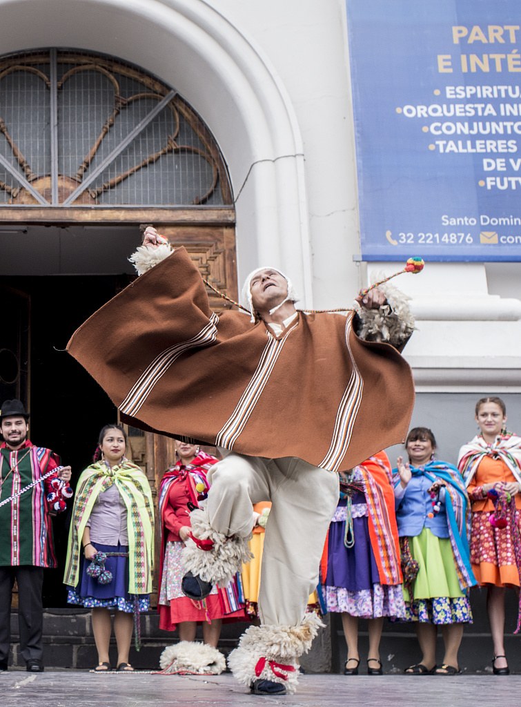 An older man in a brown striped poncho leaps high in front of a church while llamas stand nearby and an ensemble in colorful traditional dress watches.