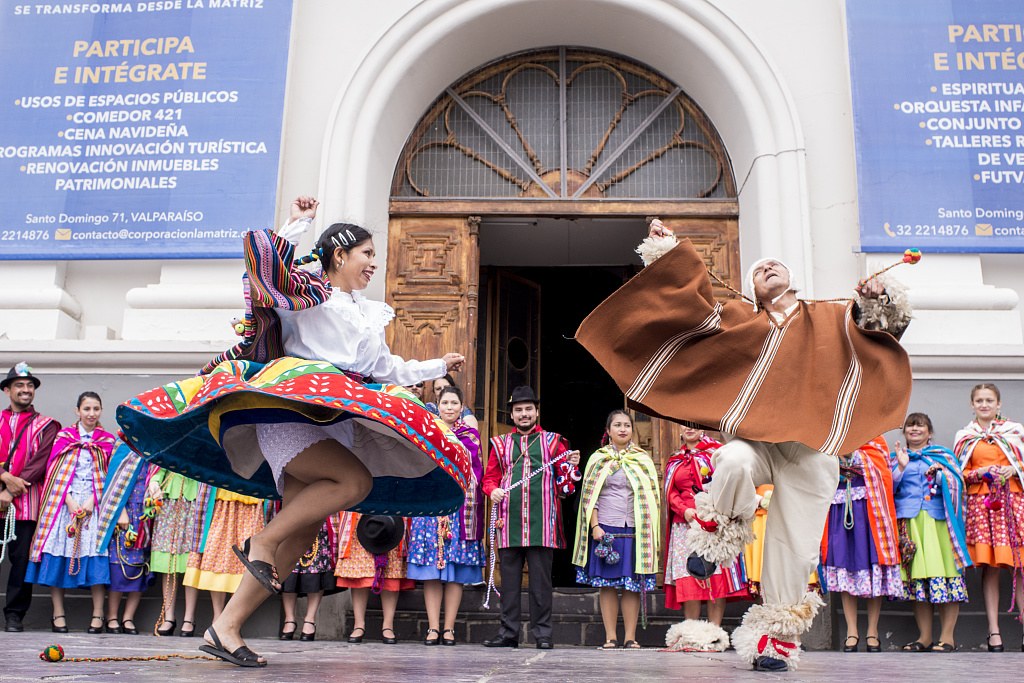 Two dancers in bright traditional Chilean costume perform cueca in front of a church, surrounded by costumed community members and onlookers.