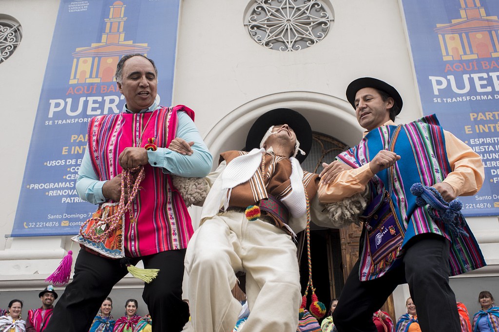 Three dancers in traditional Andean dress perform folk choreography on the steps of a white colonial church in Valparaíso, with spectators gathered below.