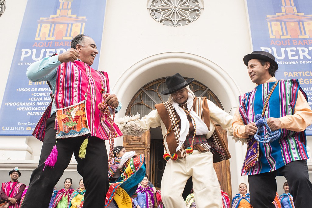 Three men in bright woven ponchos and traditional dress perform folk dance with raised arms in front of a white colonial church in Valparaiso.
