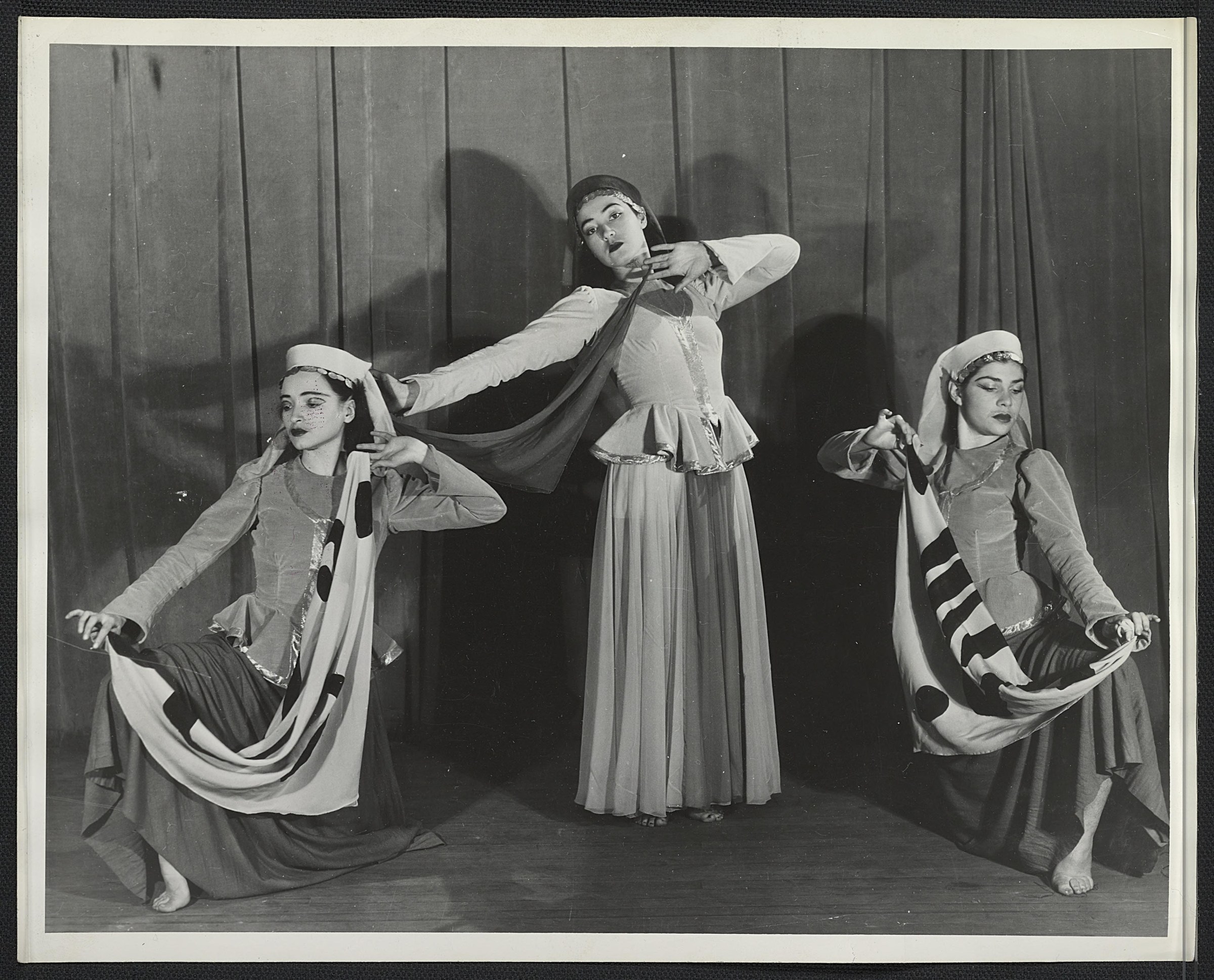 Three young dancers in folk costume on a stage: one elevated in center with raised arm, two kneeling on either side holding flowing fabric.